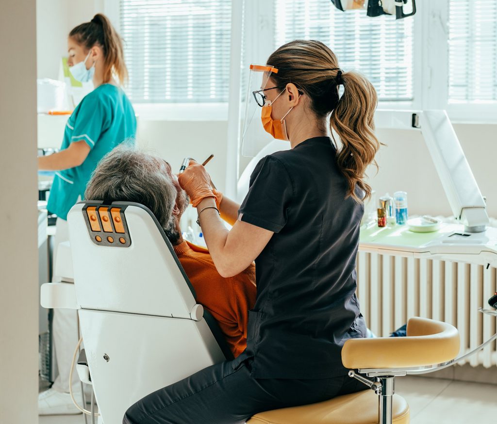 Terms and Conditions 1 Dentist in face shield treating an older male patient in the chair while a dental assistant preps instruments